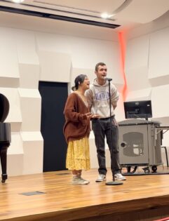 Photo of two people singing into a mic with an orange streamer behind them.